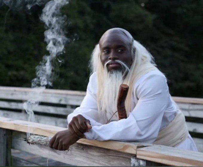 A cosplay photo of a Black man with long white hair and beard, dressed in traditional-style white robes holding a staff, with smoke rising nearby.