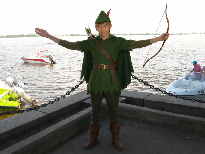 A young man dressed as Robin Hood, complete with green tunic, feathered cap, and drawn bow, poses on a pier beside boats labeled "P28" in a "Robinhood Crypto" branded promotional post announcing "$OGSM" token trading availability on Uniswap via Robinhood Chain.