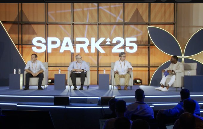 Four men sit on a stage in white armchairs at the "SPARK 25" conference, participating in a panel discussion in front of an audience, with one speaker on the right gesturing as he addresses the group. The setting and formal panel arrangement align with the post's focus on a discussion about banks transitioning to on-chain operations.