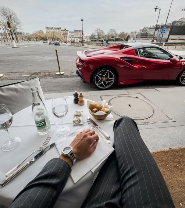 A first-person view of a person in a pinstripe suit dining at a sidewalk café, with a red Ferrari sports car parked on the Parisian street just beyond the table.
