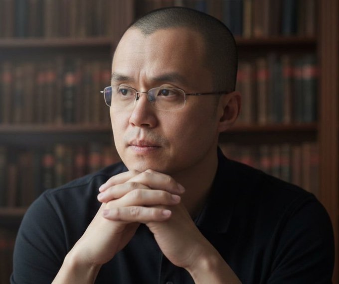 A contemplative man with glasses, shaved head, and clasped hands gazes thoughtfully in front of a bookshelf backdrop, reflecting the thoughtful, community-focused tone of CZ's message about engaging positively with crypto communities and meme culture around BNB.