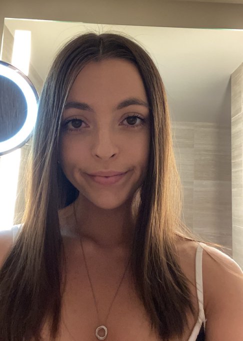 A selfie of a young woman with long brown hair wearing a white top and a pendant necklace, taken in front of a ring light in what appears to be a bathroom.
