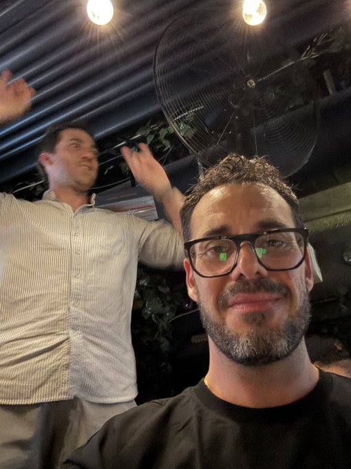 A close-up selfie shows a bearded man with glasses smiling in the foreground, while another man in a striped shirt gestures with raised hands in the background near a large fan.