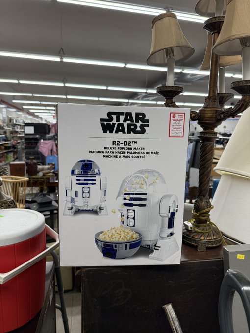 A Star Wars R2-D2 Deluxe Popcorn Maker box displayed on a surface in what appears to be a thrift store, surrounded by lamps and other merchandise.