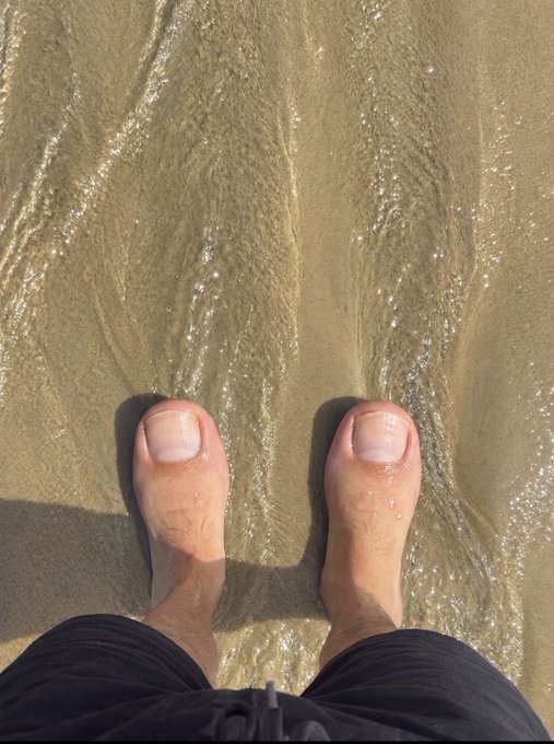 A first-person view of bare feet standing in shallow ocean water as waves wash over the sandy beach.