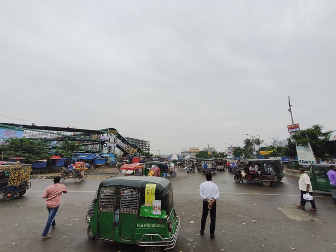 A street scene showing multiple green auto-rickshaws (CNGs) and pedestrians on a road, with construction or demolition visible on a building to the left, under an overcast sky.