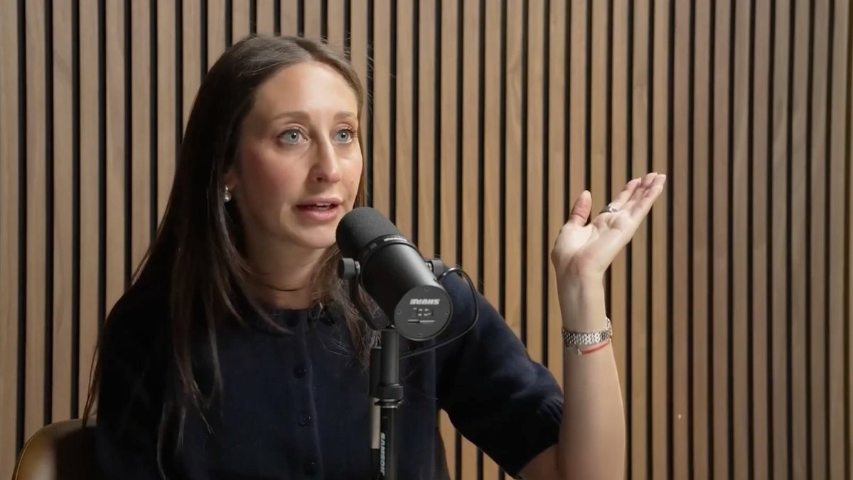 A woman with long dark hair speaking into a Shure SM7B microphone while gesturing with her hand, seated in a wood-paneled podcast or interview studio.