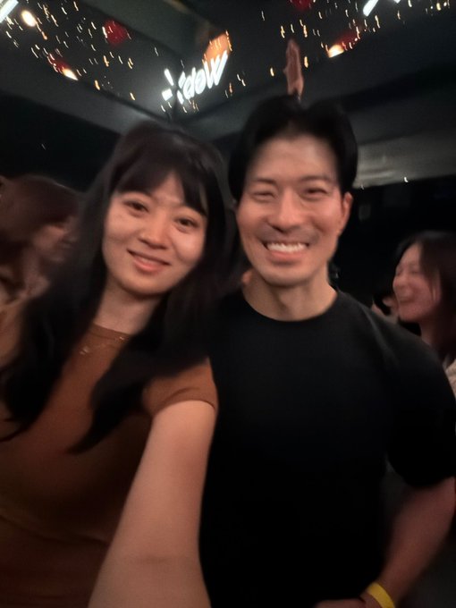 A smiling couple takes a selfie together at what appears to be a festive event or club with lights visible in the background.