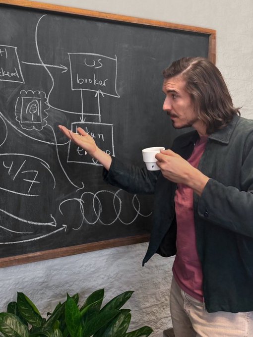 A man with long hair holds a coffee cup and gestures toward a chalkboard displaying a diagram with labels like 'US broker' and 'plan to IPO'.