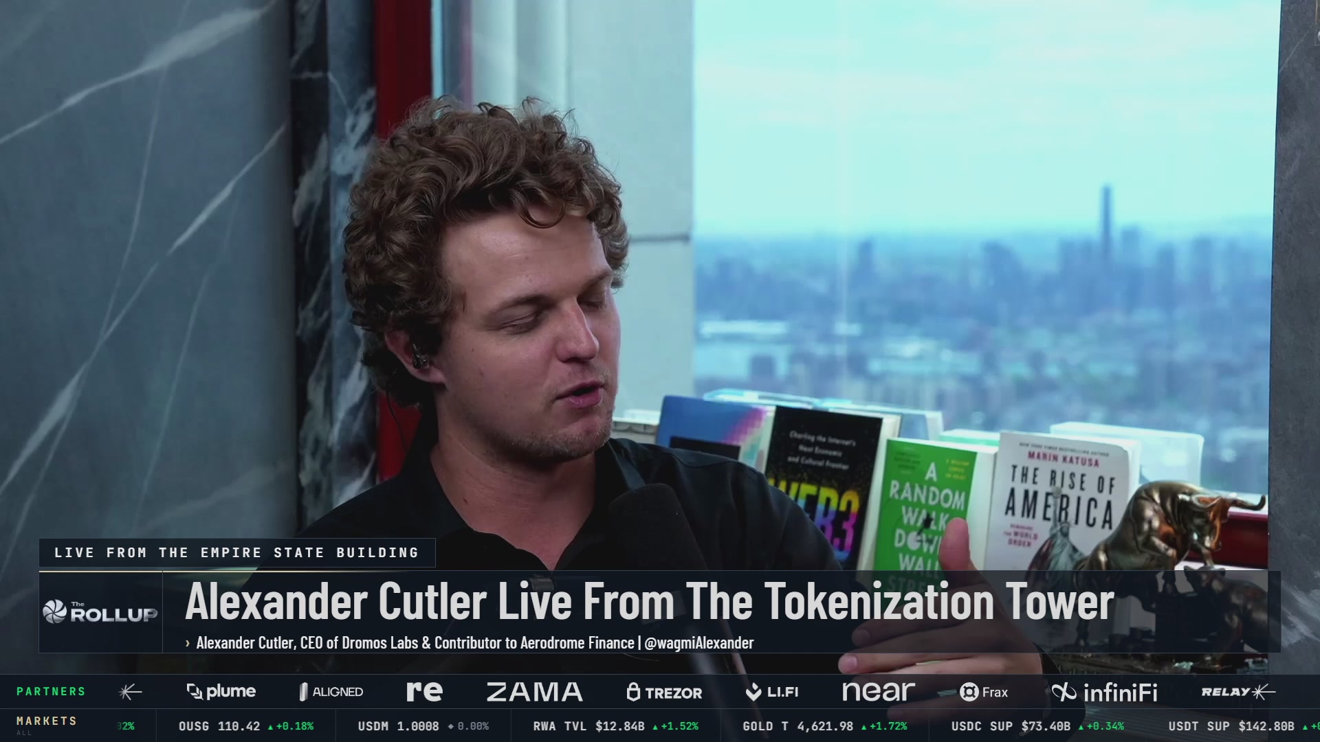 A man with curly hair, dressed casually, reads a book titled "The Bitcoin Ripple" while seated at a desk or table, with the text "ALEXANDER CUTLER Live From The Bitcoin Ripple" and "Ripple" displayed on a banner above him.