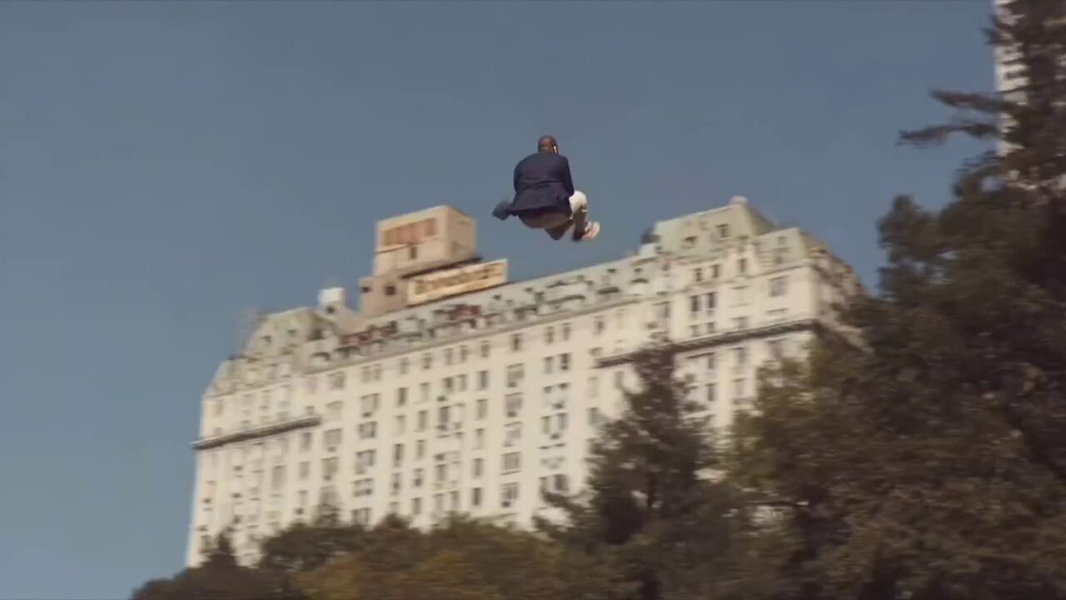 In the image, a man wearing a suit is seen flying through the air above an urban landscape with buildings of various sizes. One building has a sign that reads "The Dog will see you in the chat", which adds a humorous element to the scene. The man appears to be jumping over this building while holding onto a box, creating a sense of motion and excitement.