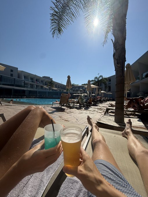 Two people relax on sun loungers by a resort pool, toasting with a blue cocktail and a glass of beer under a bright sunny sky framed by a palm tree. The leisurely vacation scene embodies "Tokenminimizing" — savoring meaningful moments in person rather than sharing them across countless social tokens.