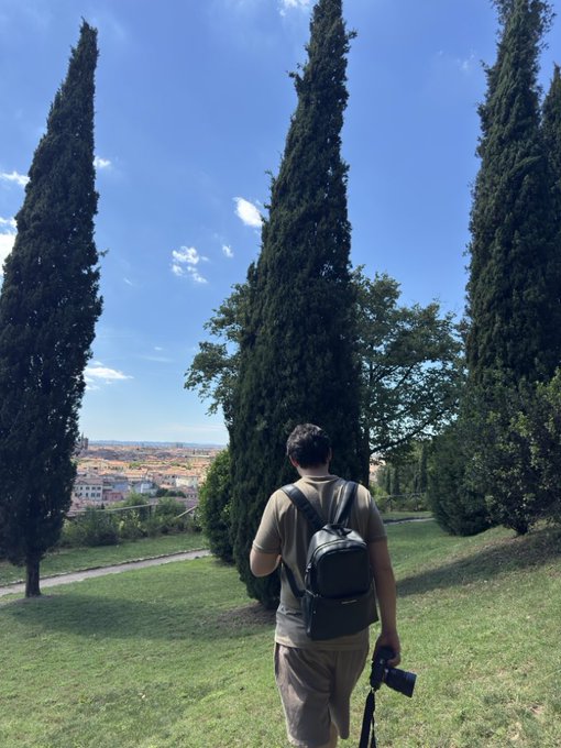 A person wearing a backpack and holding a camera stands on a grassy hill between tall cypress trees, overlooking a town under a sunny blue sky.