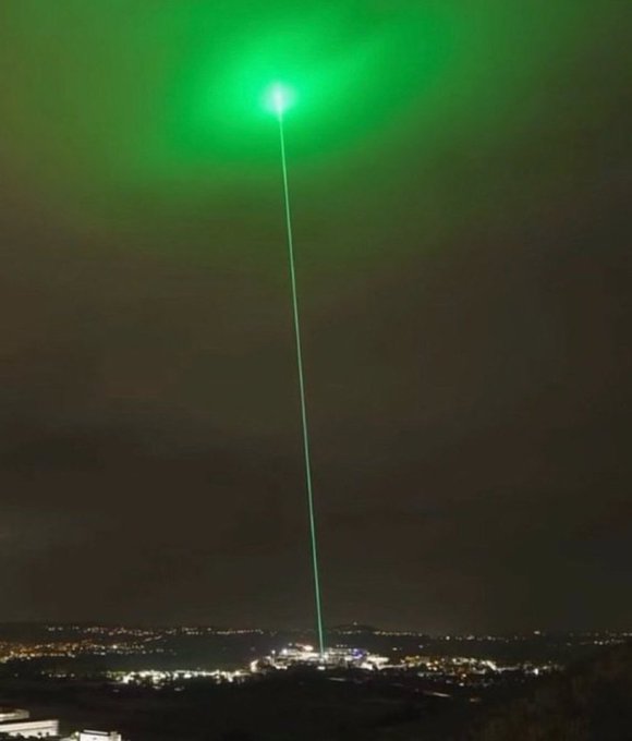 A bright green laser beam shoots into the night sky above a cityscape, illuminating the clouds with an otherworldly glow, with no visible text or signage. The dramatic "CLAIM IT." caption reinforces the image's bold, seize-the-moment energy, as if the laser itself is staking a claim on the heavens.