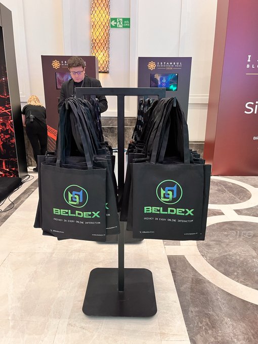 An indoor event scene with a metal display rack holding multiple black tote bags branded 'BELDEX' with the tagline 'AGENCY IN EVERY ONLINE INTERACTION'.
