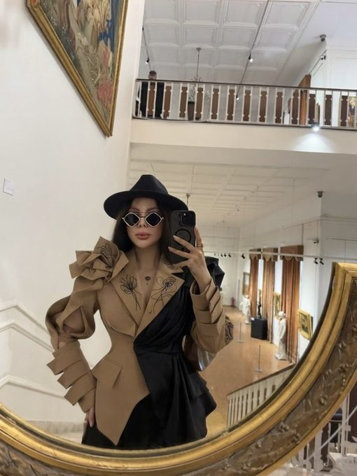 A person wearing a black hat and sunglasses takes a mirror selfie in a brown and black structured blazer in a building with a balcony.