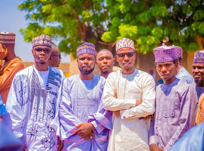 A group of men dressed in traditional Nigerian attire, including embroidered caps and flowing robes, pose together outdoors under lush green trees. The image complements the post by visually representing the community of supporters standing behind Oga Sunusi, emphasizing unity and collective commitment to the project's success.
