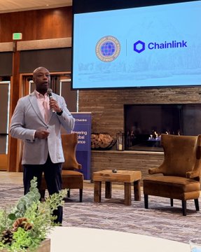 A man in a light gray suit speaks into a microphone at an event, with a screen behind him showing the Chainlink logo.