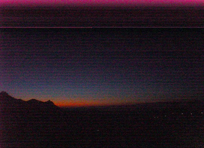 A grainy, low-light photograph captures a sunset with a vibrant orange glow along the horizon beneath dark silhouetted mountains and a twilight sky.