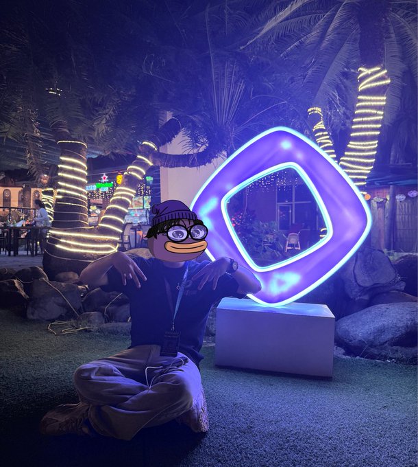 A person wearing a Pepe the Frog mask poses next to a glowing purple and blue geometric sculpture at night, with palm trees wrapped in lights in the background.