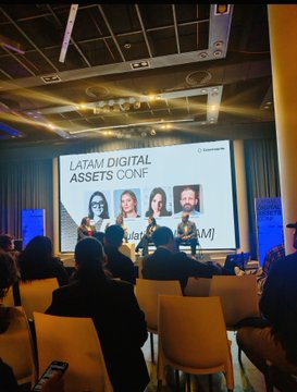 Indoor conference photo showing a panel discussion on stage at 'LATAM Digital Assets Conf' with multiple speakers seated at a table while audience members watch from the foreground.