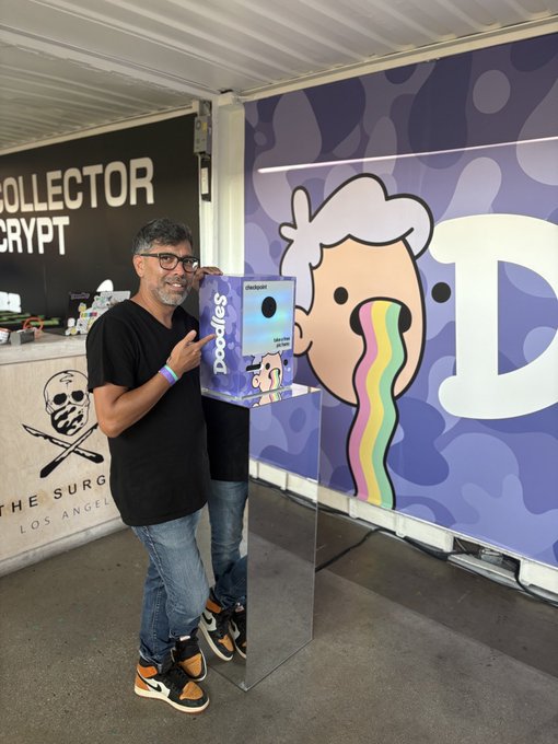 A man wearing glasses and a black shirt stands at The Surgeon Los Angeles pop-up, holding a Doodles NFT collectible box next to a large Doodles character banner with a rainbow tongue.
