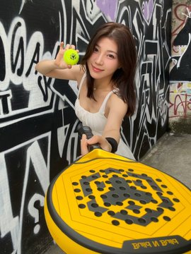 A young woman poses outdoors in front of a graffiti-covered wall, holding a yellow hexagonal workout balance pad with the text 'Train in Peace'.