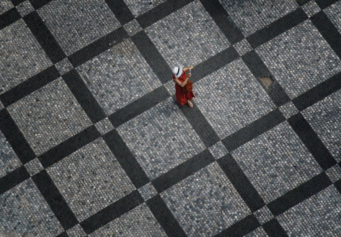 An overhead view of a person in a red and white outfit and white hat walking on a large black and gray checkered cobblestone plaza.