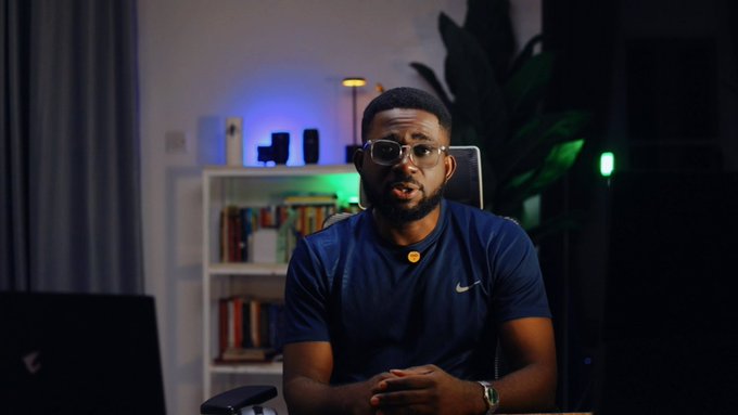 A man wearing glasses and a dark blue Nike t-shirt sits in a dimly lit room with a bookshelf and a plant illuminated by green and blue lights in the background.