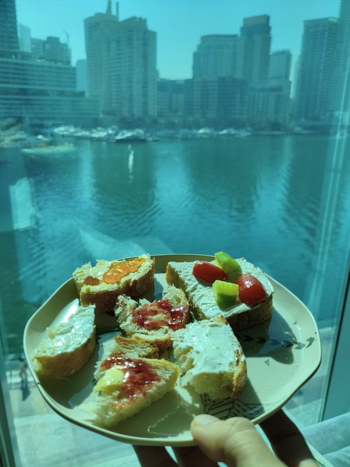 A hand holds a plate of jam-topped bread slices and cream cheese with tomatoes in front of a window overlooking a harbor and city skyline.