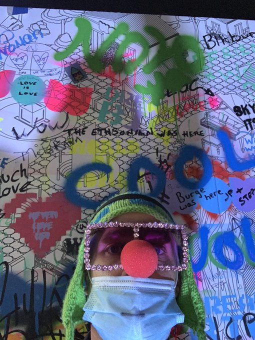 A person wearing a green knit hat, decorative sunglasses, a red clown nose, and a surgical mask poses in front of a graffiti-covered wall with messages like 'THE ETHBORN WAS HERE' and 'LOVE IS LOVE'.