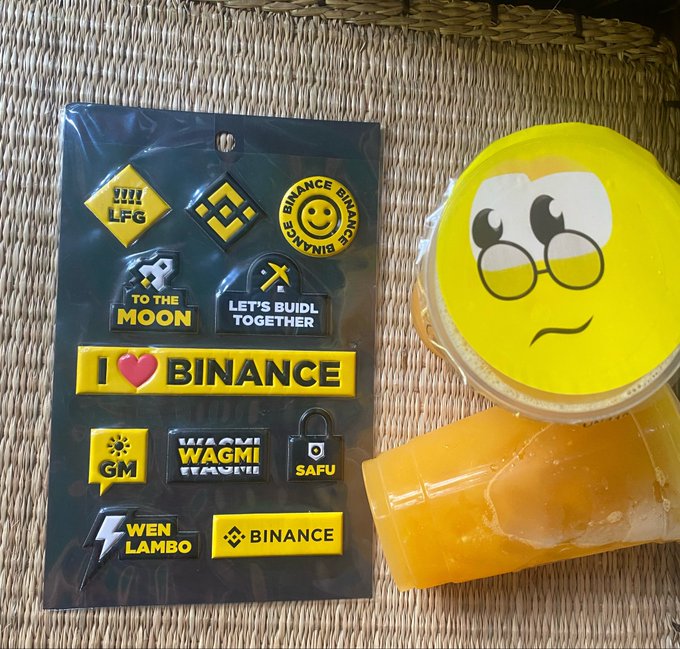 A sheet of Binance-themed stickers with slogans like 'TO THE MOON', 'LET'S BUILD TOGETHER', 'I ❤ BINANCE', 'WAGMI', 'SAFU', and 'WEN LAMBO' sits on a woven mat next to an orange drink with a worried-face emoji lid.