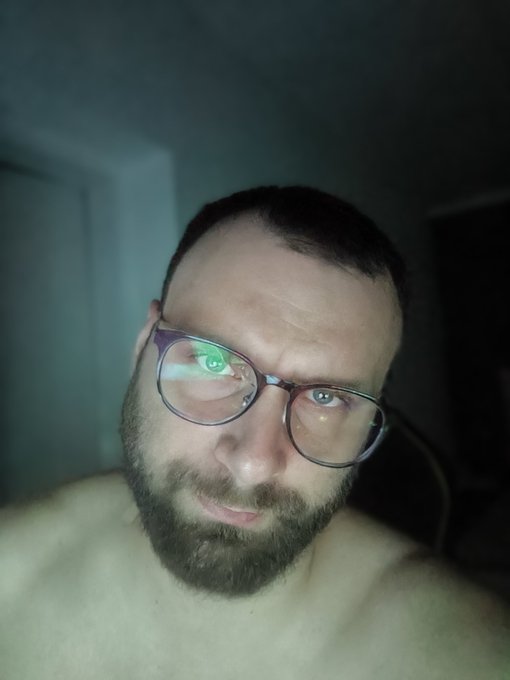 A shirtless man with glasses and a full beard looking into the camera, with a green light reflection visible in one of his lenses.
