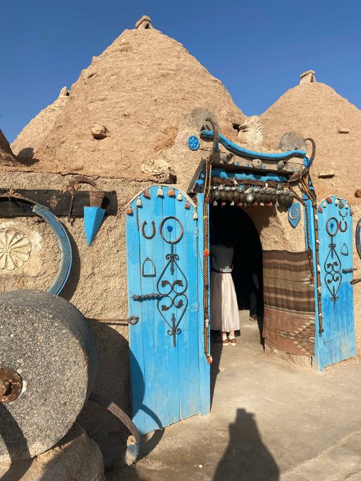 The image shows an old blue door with intricate designs, possibly made of stone or wood, standing out against a clear sky. The door has two windows above it, one larger than the other, adding character to its appearance. A person can be seen in front of the door, likely entering or leaving the building.