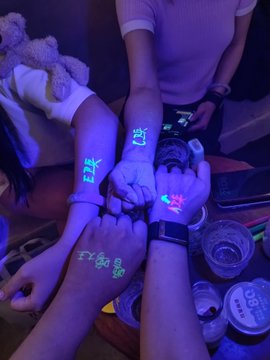 Several people's arms and hands are gathered together under purple UV lighting, with Korean text written in glow-in-the-dark ink on their skin, suggesting a fan event or party.