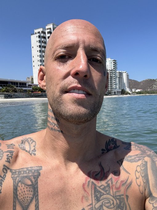 A bald, tattooed man takes a selfie at the beach with high-rise buildings and mountains visible across the water, capturing a relaxed day in a coastal resort town.