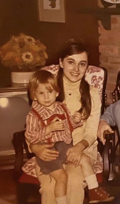 A vintage, sepia-toned photograph shows a smiling woman with long dark hair sitting in a floral-patterned chair, holding a young child with light hair on her lap; the background includes a vase of flowers on a side table.
