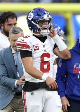 New York Giants quarterback Jaxson Dart (#6) stands on the sideline, wearing his white away jersey with the "C" captain's patch and NY helmet, as he appears to be receiving attention from team staff following his potentially season-ending knee injury.