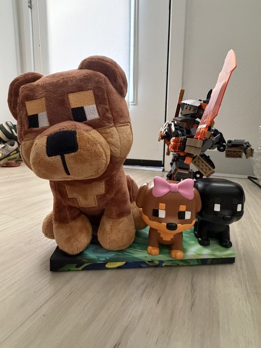 A collection of figurines on a Minecraft-themed base, including a large Minecraft bear plush, a Lego-style figure with a sword, a small brown dog figure with a pink bow, and a black Minecraft dog figure.
