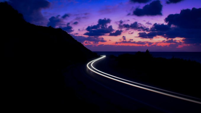 A winding coastal road curves through a darkened landscape under a vibrant sunset sky of purple, pink, and deep blue clouds, with long-exposure light trails tracing the curve of the pavement. The image evokes a sense of journey and transition, fittingly complementing the artist's grateful post celebrating the sale of their photographic work "Lineas" to collector @tokioflo.