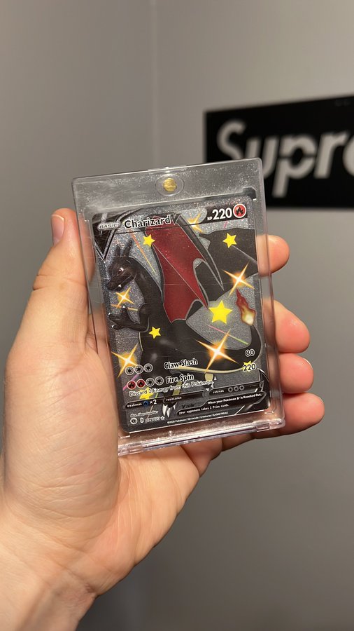A hand holds a Charizard Pokémon trading card encased in a protective plastic sleeve, with a partially visible Supreme sign in the background.