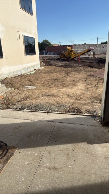 A construction site next to a beige building, showing a dirt lot, gravel, a yellow piece of machinery, and orange safety cones under a clear blue sky.