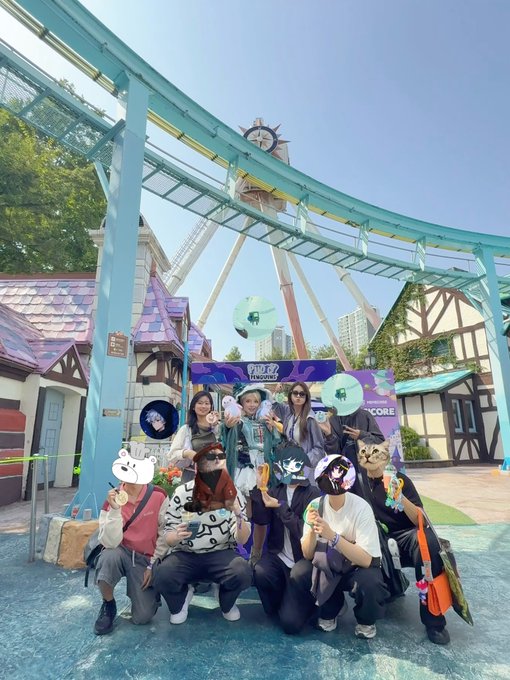 A group photo of nine people posing at an amusement park in front of a clock tower structure; several people have anime character emojis covering their faces, and a sign reading '外星人工作室' (Alien Studio) is visible.
