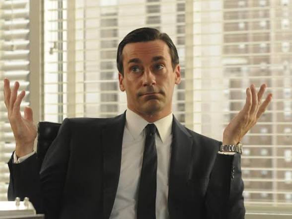 A man in a dark suit and tie sits with both hands raised palms-up in a shrugging gesture, with venetian blinds visible behind him.