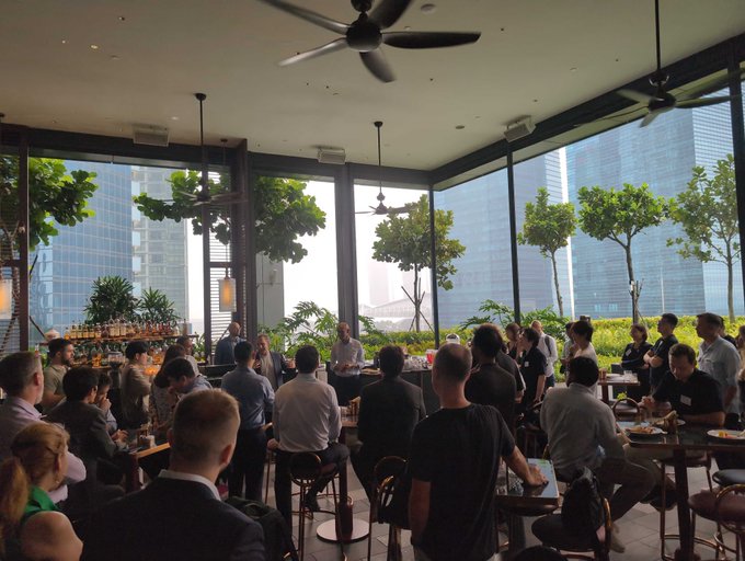 An indoor photograph of a crowded rooftop bar or restaurant with large windows showing a city skyline, where a speaker is addressing patrons at a networking or presentation event.