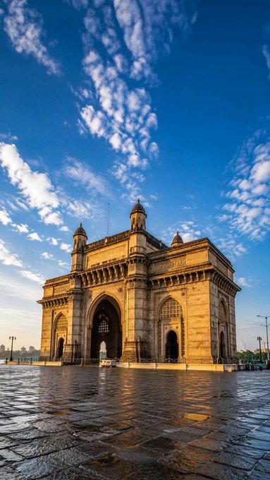 The Gateway of India monument in Mumbai stands against a vivid blue sky with scattered clouds, its yellow stone facade glowing in the warm sunlight.