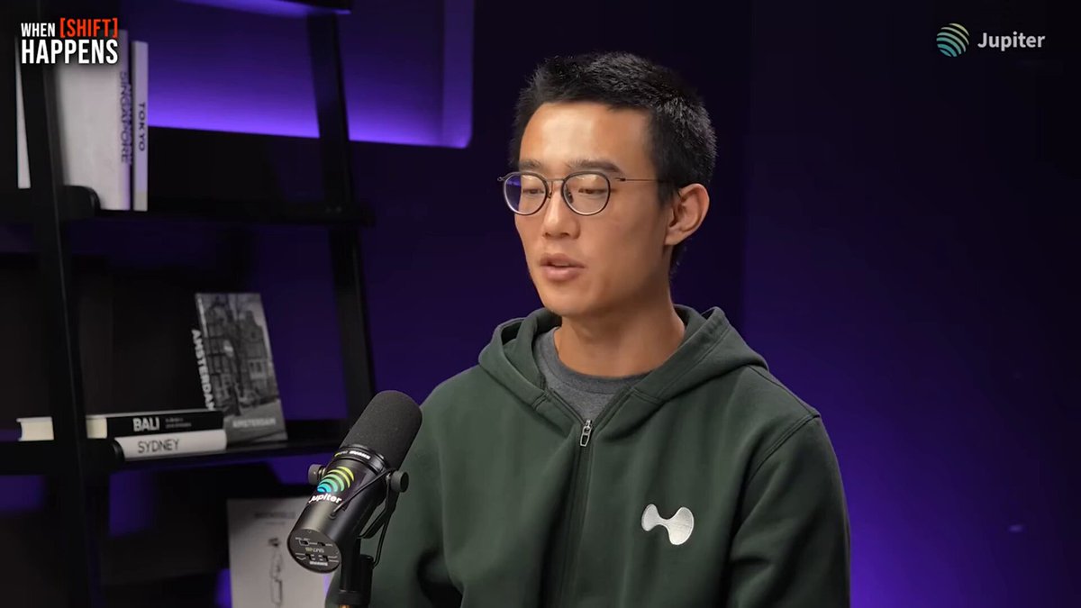 The image features a man wearing glasses and a green hoodie standing in front of a microphone, giving him an air of authority or expertise as he speaks into it. The background includes bookshelves filled with various books, suggesting that this might be a podcast recording session taking place at a library or a similar setting. 

The image also contains the text "Sports outcome markets, now live on Hyperliquid." This could indicate that the man speaking is discussing sports-related topics and how they are being covered by online platforms like Hyperliquid. The presence of bookshelves in the background further supports this idea, as libraries or bookshops often host podcasts or discussions related to various subjects, including sports events and their impact on society.