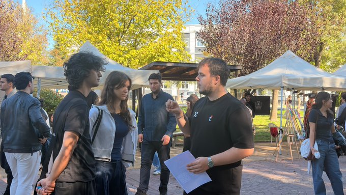 People stand and talk outdoors near white event tents and trees, while a man in a black shirt gestures with papers in his hand.