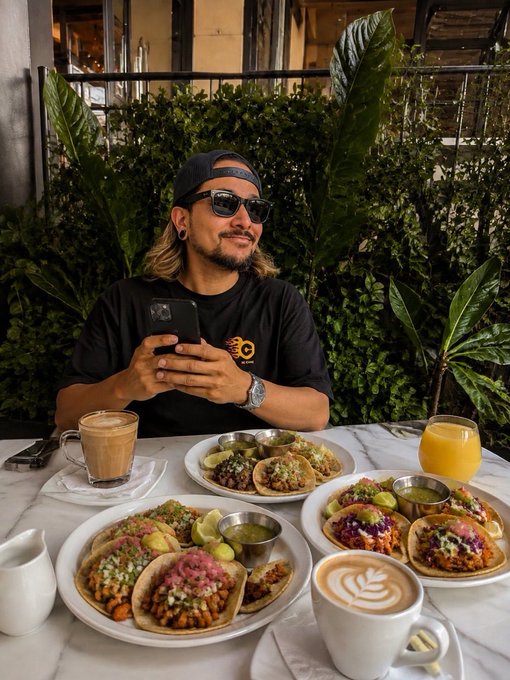 A man wearing sunglasses and a backwards cap smiles while checking his phone at a restaurant, surrounded by three plates of colorful tacos and coffee drinks. The spread of tacos pairs perfectly with the "Gm & Happy Taco Tuesday" caption, showcasing a vibrant and satisfying brunch scene.