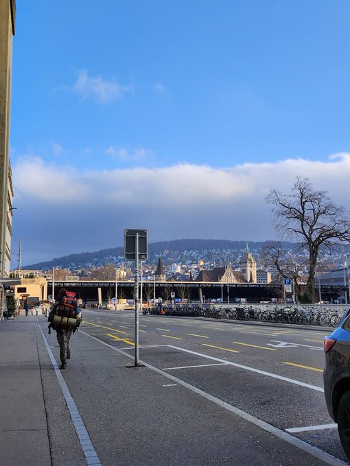 A street view showing a person with a large backpack walking along a road with bicycles parked, overlooking a city with hills in the distance under a partly cloudy blue sky.